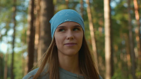 a woman with a blue headscarf and a warm smile walks through a sunlit forest, her dimples visible as she gazes contentedly around, sunlight filters gently through tall trees