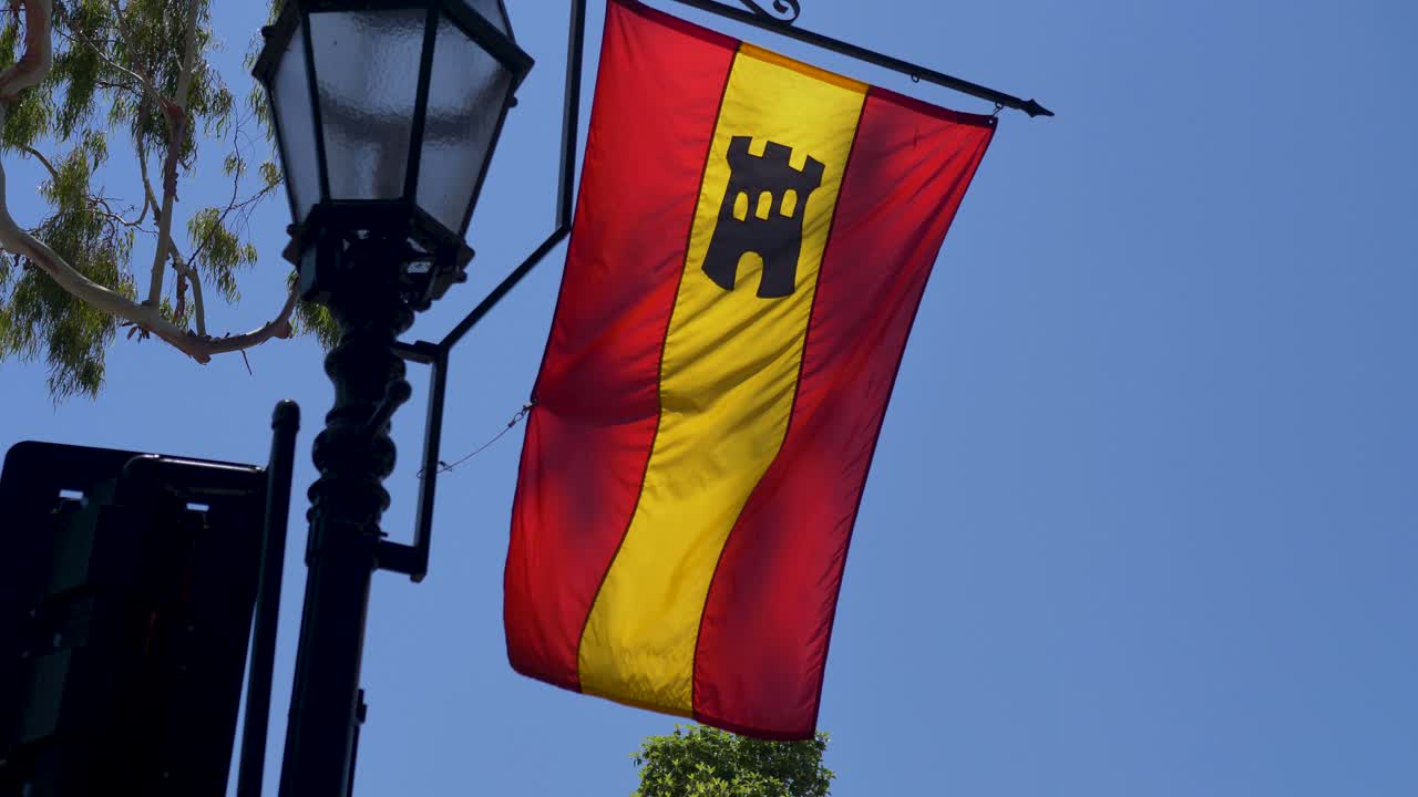 A red and yellow flag with a black castle symbol hangs from a lamppost against a clear blue sky.