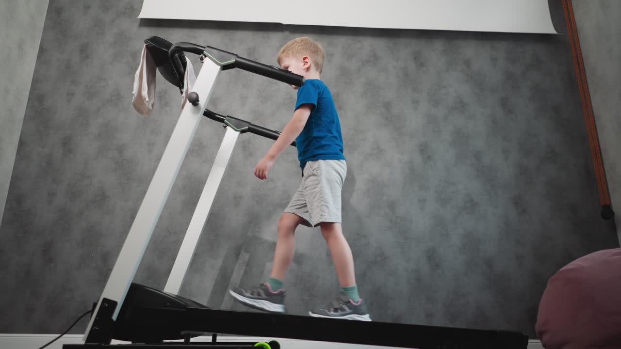 young boy walking steadily on treadmill in workout session focused on movement and balance with towel hanging from console against grey wall and wooden floor in modern indoor home environment