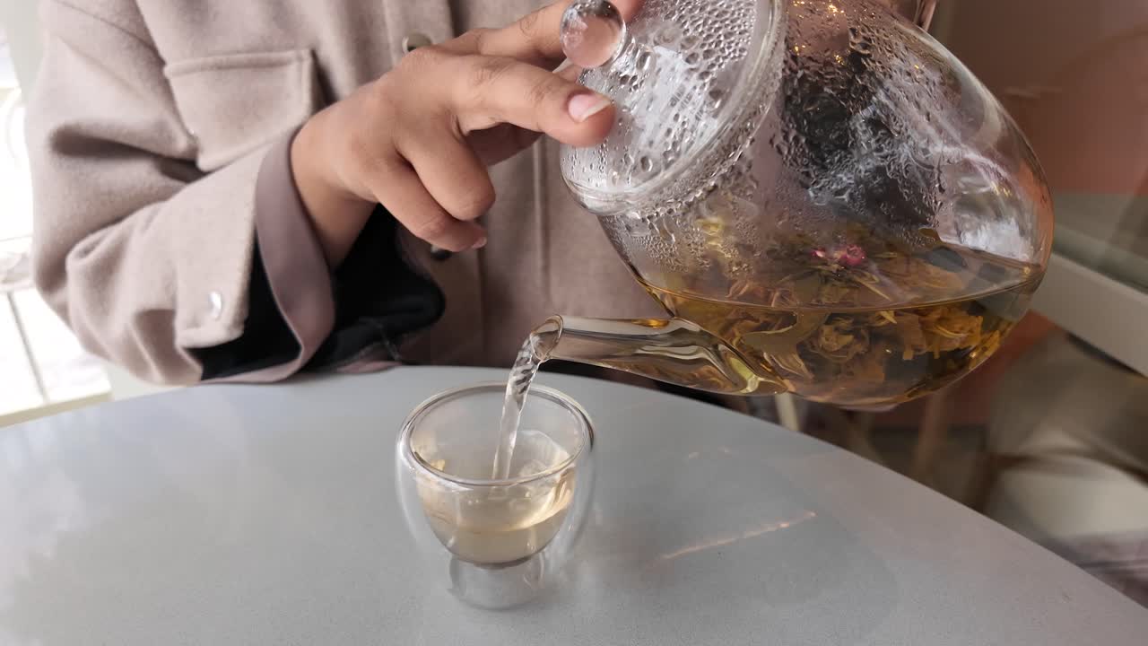 Primo piano di mani che versano tè ai fiori da una teiera di vetro a una tazza
