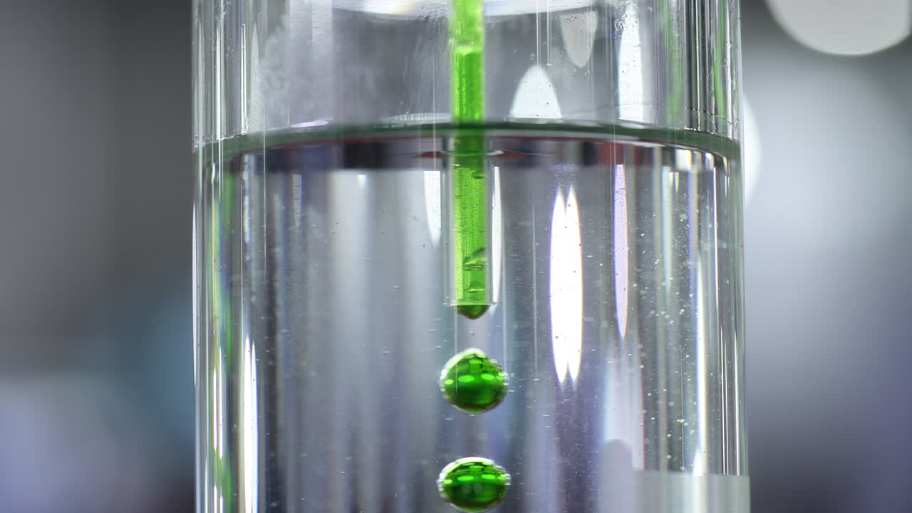 Green Liquid Drops Falling Through Clear Water in Glass Cylinder