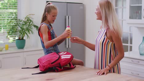 Madre-Caucásica-Sonriente-En-La-Cocina-Con-Su-Hija-Preparando-La-Mochila-Escolar,-Empacando-Jugo-Y-Hablando