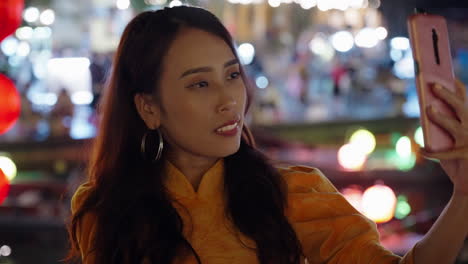 Woman-in-yellow-Ao-Dai-taking-selfies-amidst-Hoi-An's-vibrant-nightlife,-colorful-bokeh-lights