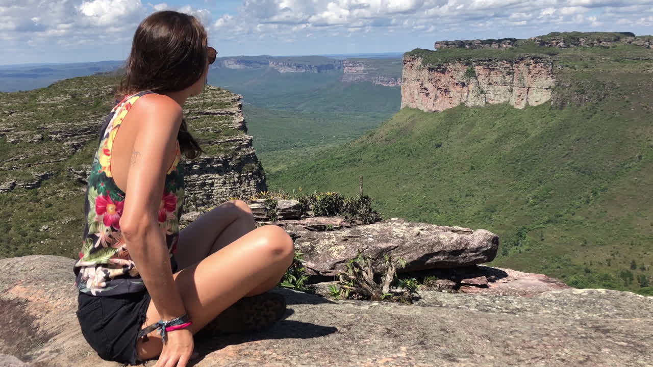 Young woman enters frame and sits down to calmly look at beautiful mountain landscape, Brazil, Slowmotion