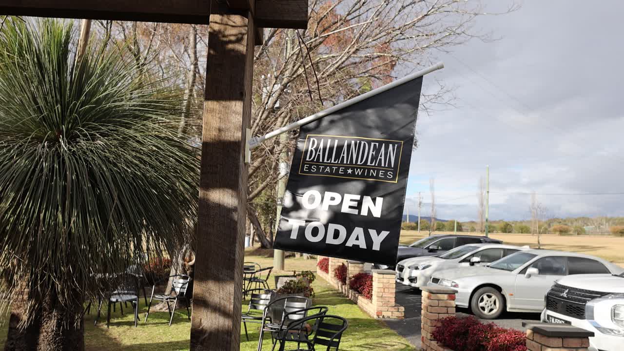 Black winery sign sways in wind near parked cars, outdoor seating, and native plants, daylight