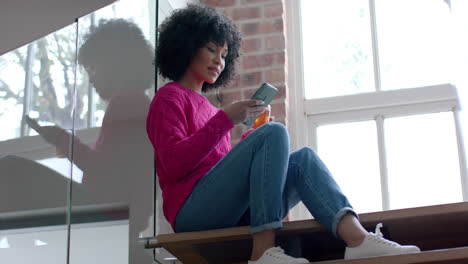 mujer biracial bebiendo té y usando teléfono inteligente en casa, cámara lenta