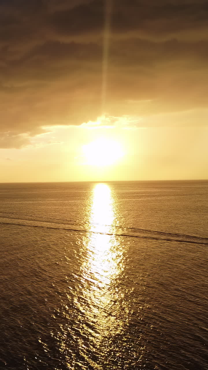 Luftfoto fra drone af en dramatisk gylden solnedgang over havet langs den mexicanske kystlinje. En lille båd krydser det rolige hav og efterlader en lang kølvandsstribe under farverige skyer og varmt aftenlys. Vertikal.