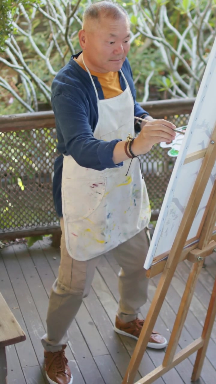 Vertical video: Facing canvas, Asian man in apron dipping brush, refining work on deck with palette