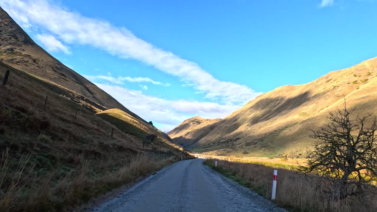Vehicle travels scenic gravel road, sunlit mountains, clear sky, wide angle, steady forward motion