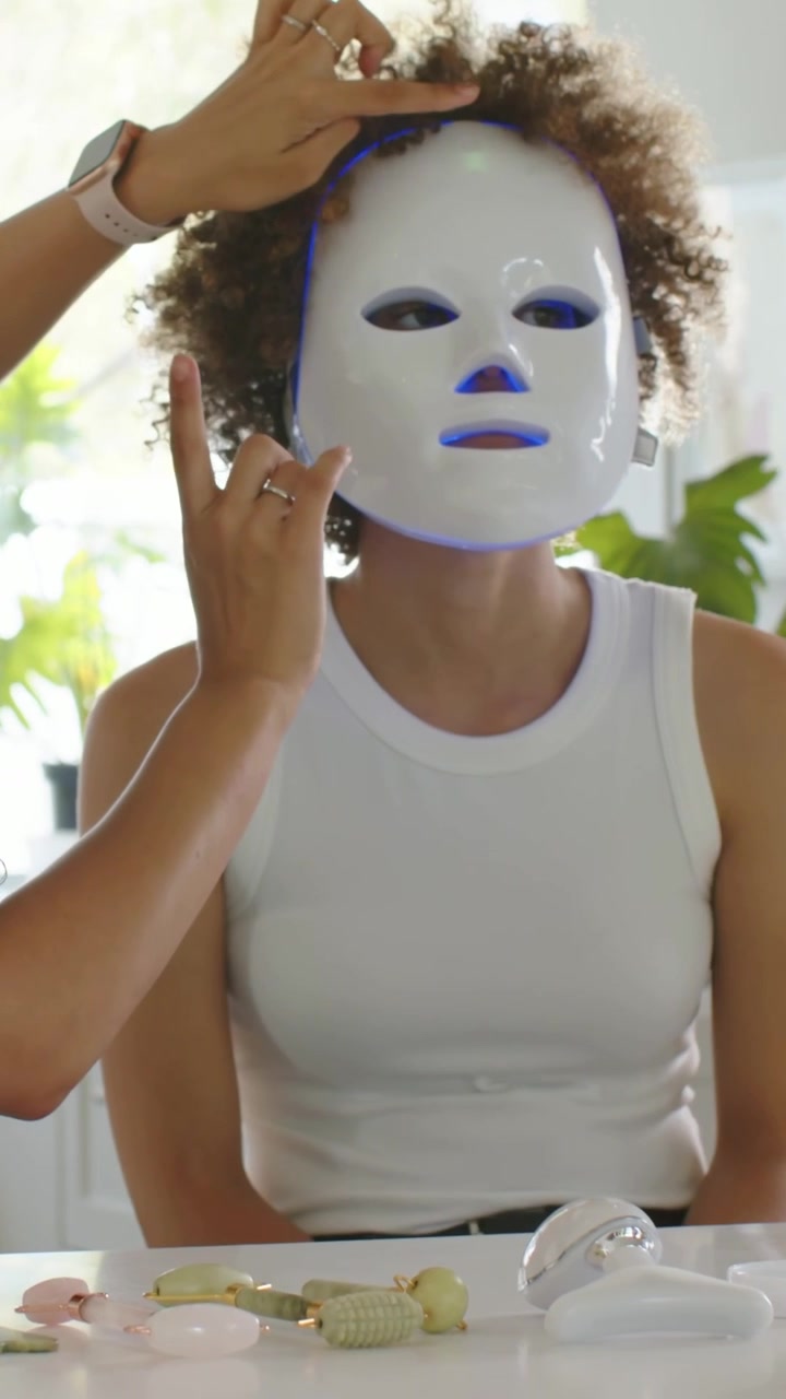 Vertikal video: Talende vært i grøn top placerer LED-maske på kunde i køkken, viser hudpleje.