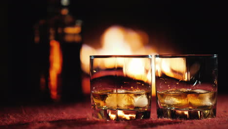 Zwei-Gläser-Whisky-Mit-Eis-Im-Hintergrund-Eine-Flasche-und-Ein-Kamin,-In-Dem-Das-Feuer-Brennt