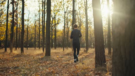 Ein-Mann-In-Schwarzer-Sportuniform-Mit-Lockigem-Haar-Und-Turnschuhen-Läuft-An-Einem-Sonnigen-Tag-Durch-Einen-Herbstlichen-Wald-Mit-Abgefallenen-Blättern.-Seitenansicht-Eines-Glücklichen-Jungen-Sportlers,-Der-An-Einem-Sonnigen-Herbstmorgen-Durch-Den-Herbstlichen-Wald-Läuft