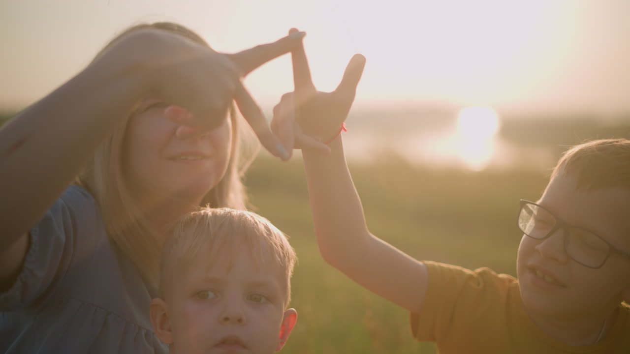 una madre con un vestido azul, sentada en la hierba con sus dos hijos pequeños durante la puesta del sol. su hijo mayor con una camisa amarilla, forman una forma lúdica con sus dedos mientras el niño más joven mira atentamente