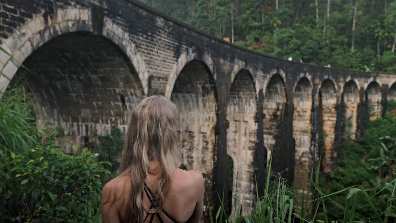 A captivating scene in Ella, Sri Lanka, where a woman walks through the lush jungle, gazing towards the iconic Nine Arches Bridge.
