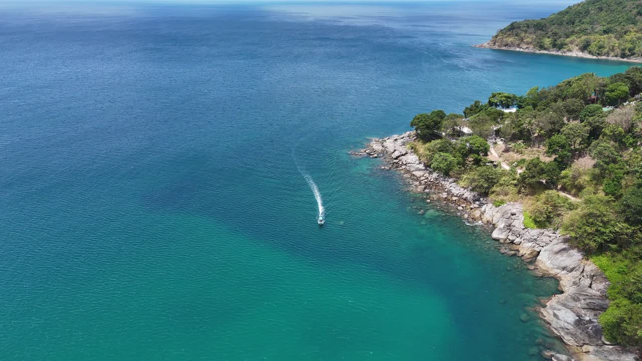 Aerial footage of a boat cruising along Phuket's rocky coastline, showcasing vibrant turquoise waters and lush greenery under clear skies