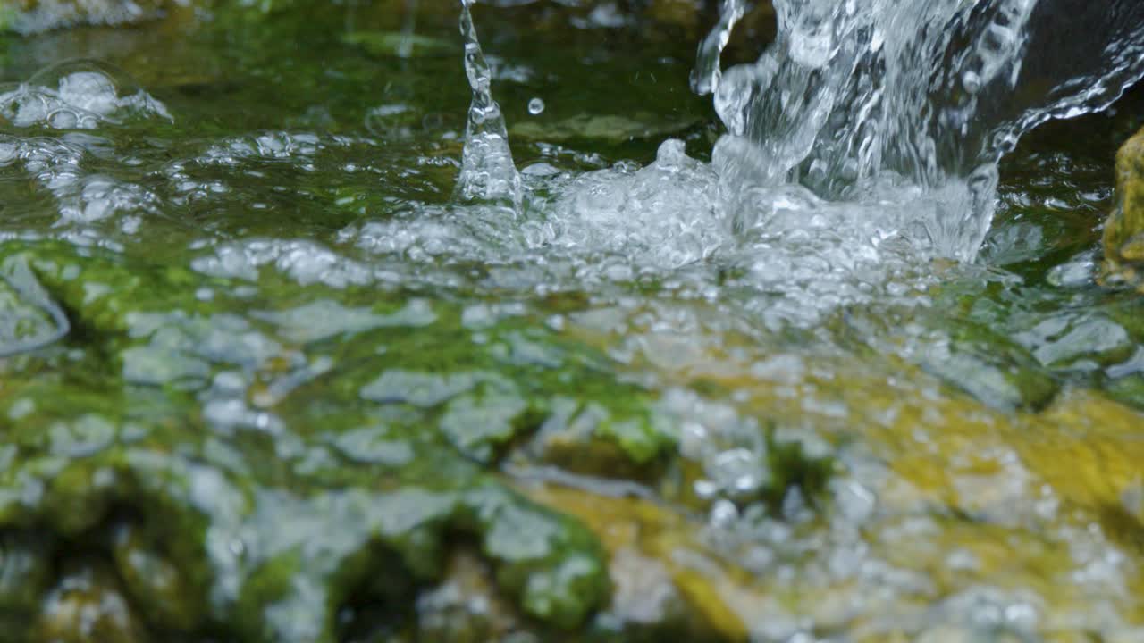 Clear water cascades over mossy rocks into a pond, creating bubbles and gentle ripples