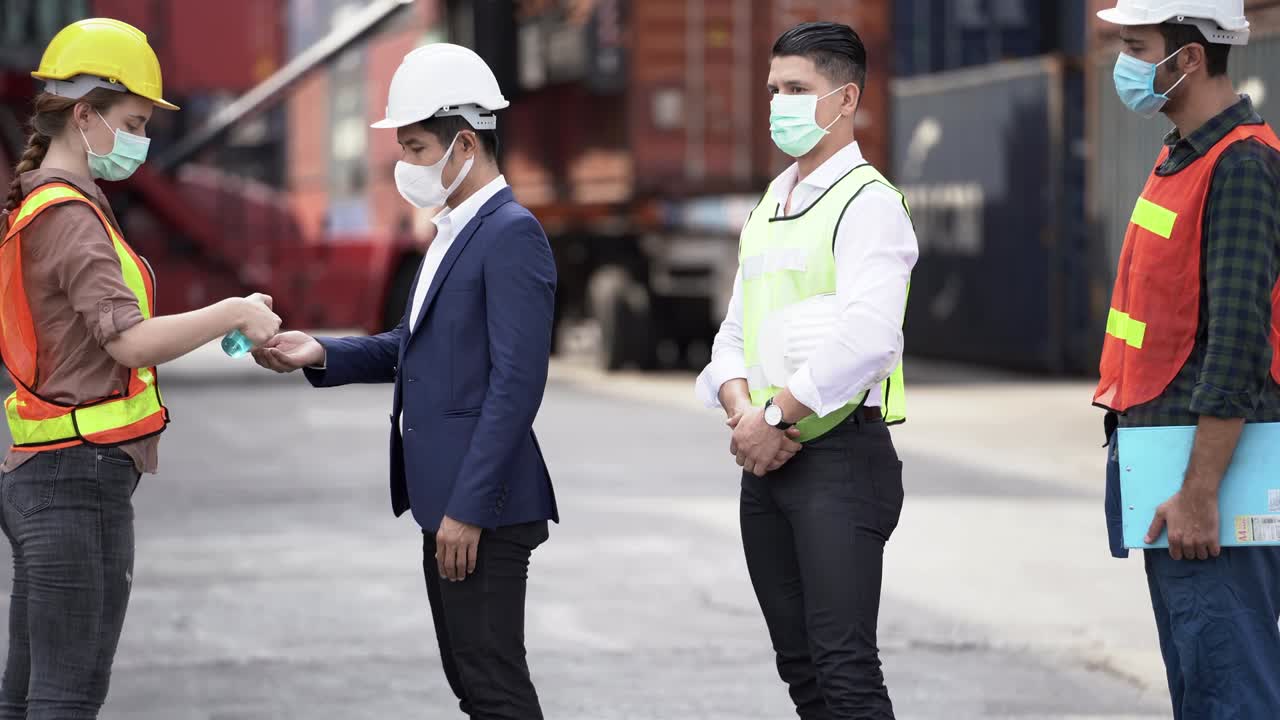 mujer trabajadora de la fábrica con una máscara médica para la cara y un vestido de seguridad usando gel de alcohol para la limpieza de manos para proteger la enfermedad del coronavirus en el almacén de la fábrica al aire libre en la gente del trabajador de pie en la cola.