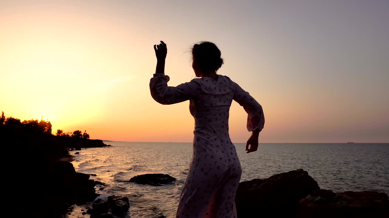 adorable chica caucásica morena en vestido claro bailando en el acantilado sobre el mar a la hora del amanecer