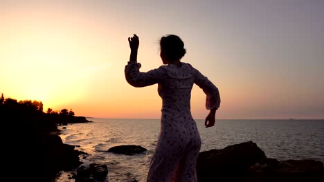 adorable caucasian brunette girl in light dress dancing at the cliff above the sea at sunrise time