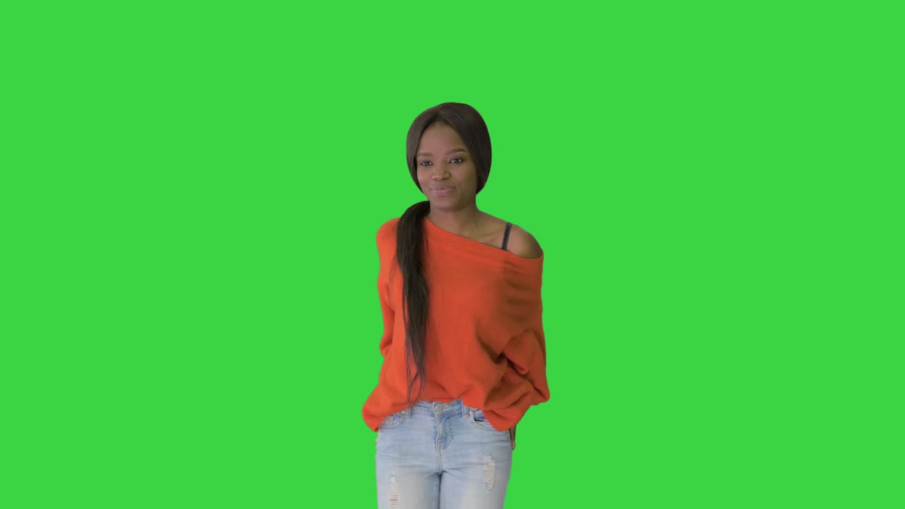 Mulher afro-americana sorridente vestindo suéter brilhante e jeans, posando com as mãos nos bolsos em uma Tela Verde, Chroma Key