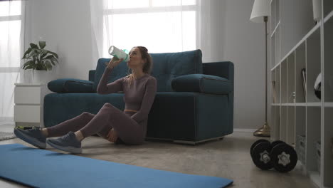 relax gym bunny is sitting on floor after home training at weekends drinking water and resting sporty woman in living room workout and wellness