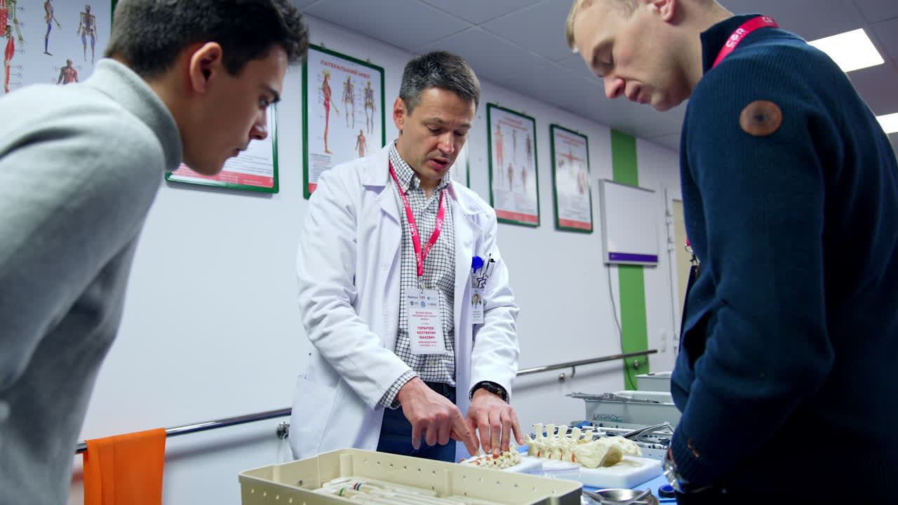 Man in white robe stands at the desk talking and pointing at the spine model. Male students listen to the speaker teaching neurosurgery. Low angle view.