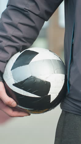 primer plano de alguien entrando en un entorno deportivo con la mano sosteniendo una pelota de fútbol negra y blanca con enfoque en la mano del jugador, mostrando el uso atlético y parte del cuerpo, fondo urbano