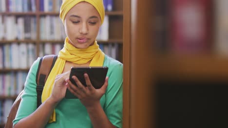 an asian female student wearing a yellow hijab leaning against bookshelves and using tablet