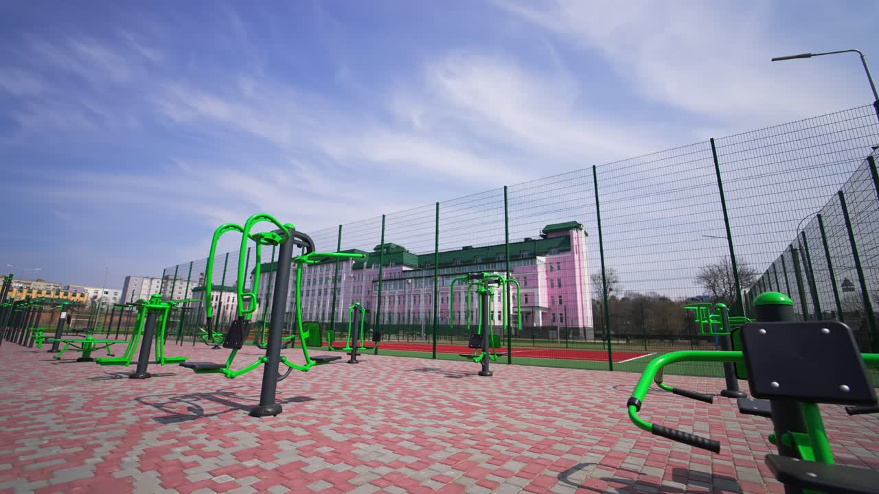 New modern sports ground with green sports horizontal bars surrounded by high metal fence. Red running tracks, basketball ground and big pink building at backdrop.