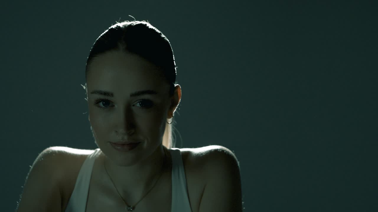 Closeup of a fit woman cycling indoors in a dark studio, smiling softly at the camera as cinematic light glows behind her, highlighting her shoulders in smooth motion from right to left