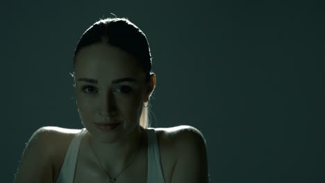 closeup of a fit woman cycling indoors in a dark studio, smiling softly at the camera as cinematic light glows behind her, highlighting her shoulders in smooth motion from right to left