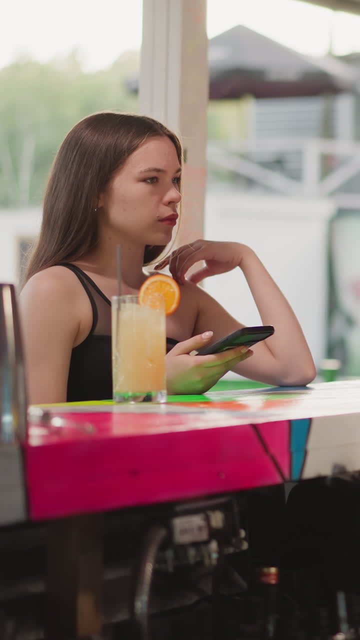 Customer waits for bill from barman in cafe. Young woman with cocktail and phone opposite black man working on computer in bar. Lady buys drink