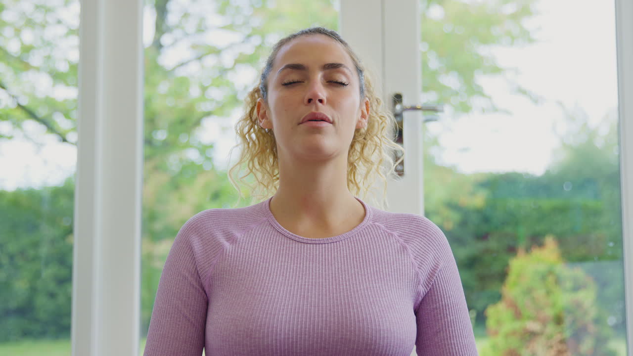 Portrait Of Smiling Young Woman Sitting On Mat At Home In Yoga Position Meditating