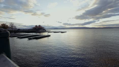 View of distant sunset over waterfront railing, showing the foreground boardwalk, boathouse, and distant mountains.