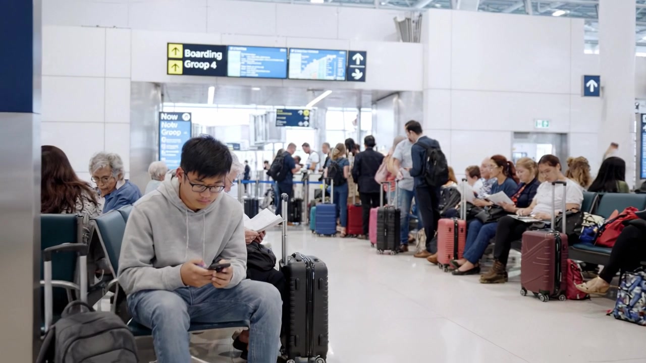 Pelancong Berkumpul di Area Boarding Bandara, Menunggu Keberangkatan dan Memeriksa Waktu di Tengah Kerumunan Penumpang dengan Bagasi dan Layar Informasi Penerbangan