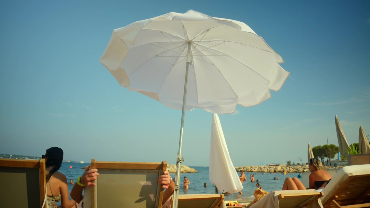 Persone che si rilassano su lettini e nuotano nel mare azzurro e limpido sotto un ombrellone bianco su una spiaggia estiva soleggiata.