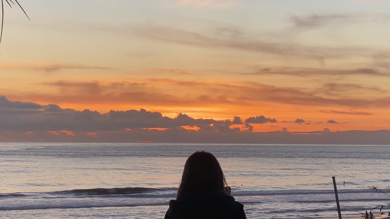 una turista viajera en la playa de carcavelos toma fotos de pequeñas olas rompiendo, una turista parada frente a una enorme puesta de sol sobre el mar en el día soleado