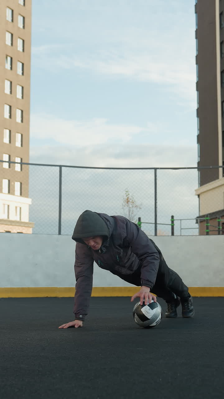 Athlete performing push ups on outdoor sport arena with alternating hand placement on soccer ball, showcasing coordination, with goal post and residential buildings in urban background