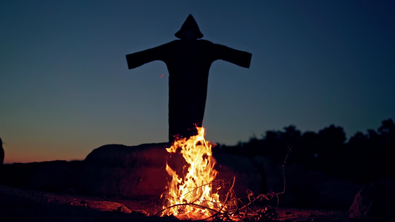 Scary dark figure with hood waving arms. Bonfire burning and mysterious ghost in black costume outdoors. Halloween costume.