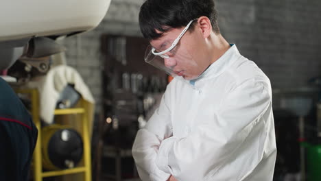 lab technician wearing face shield and lab coat stands with arms folded near car in automobile workshop, partially visible mechanic in blue uniform working in background