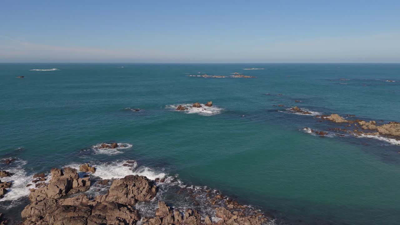 Riprese che rivelano dall'alto isole rocciose e affioramenti con onde spumeggianti che li lambiscono, ritirandosi verso la costa rocciosa e viste sull'orizzonte in una giornata limpida con cielo azzurro e sole pomeridiano.