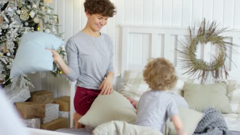 Feliz-Madre-Joven-Y-Lindos-Niños-Peleándose-Con-Almohadas-En-La-Cama-En-Una-Acogedora-Habitación-Decorada-Con-Un-árbol-De-Navidad