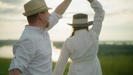 close-up of a man happily spinning his wife around on a grassy hill beside a peaceful lake at sunset. the man, dressed in a white shirt, hat, and jeans, shares a tender moment with the woman