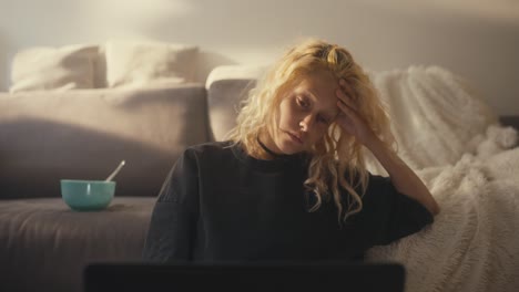 a tired blonde girl in a gray t-shirt leans on her hand and looks sadly at the laptop screen near the sofa at home