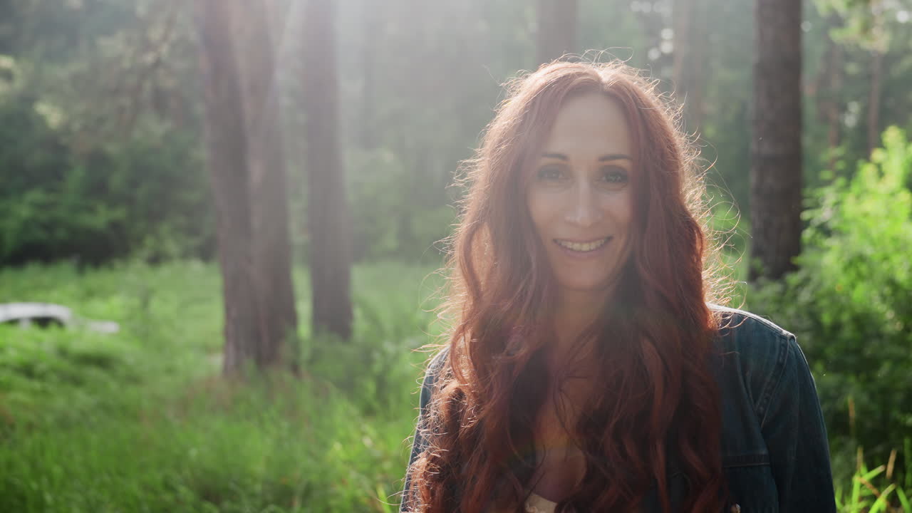 Portrait of elegant woman with long red hair smiling softly while standing near tree in sunlit forest, sunlight creating warm glow around her face as gentle breeze moves her hair, natural background