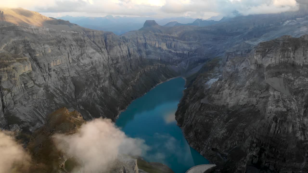 A flyover above lake Limmernsee in Glarus, Switzerland, with view of cliffs, clouds, a hydroelectric dam and reservoir after sunset