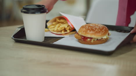 primer plano de las manos de una mujer en un vestido rosa dejando caer una bandeja negra con papas fritas, hamburguesa y café, ella ajusta la bandeja, alcanzando las papas frites, con una iluminación tenue y un fondo borroso