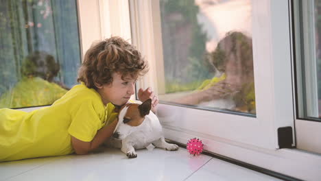 Blonder-Junge-Mit-Lockigen-Haaren,-Der-Mit-Seinem-Hund-Auf-Dem-Boden-Liegt,-Ihn-Streichelt-Und-Aus-Dem-Fenster-Schaut-2