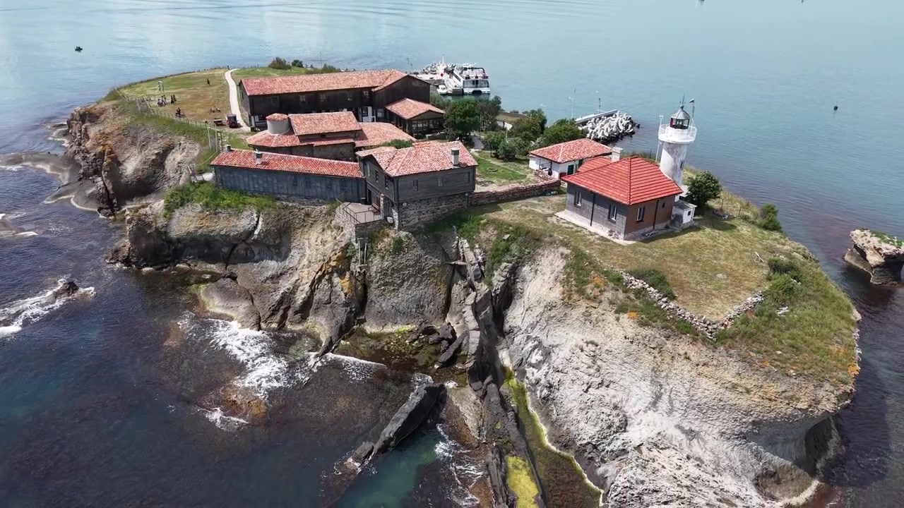Video cinematográfico con dron que captura la rocosa Península de Hel con un faro blanco, edificios con tejados rojos, serenas aguas del Báltico, suaves olas y barcos amarrados en un pequeño muelle a plena luz del día