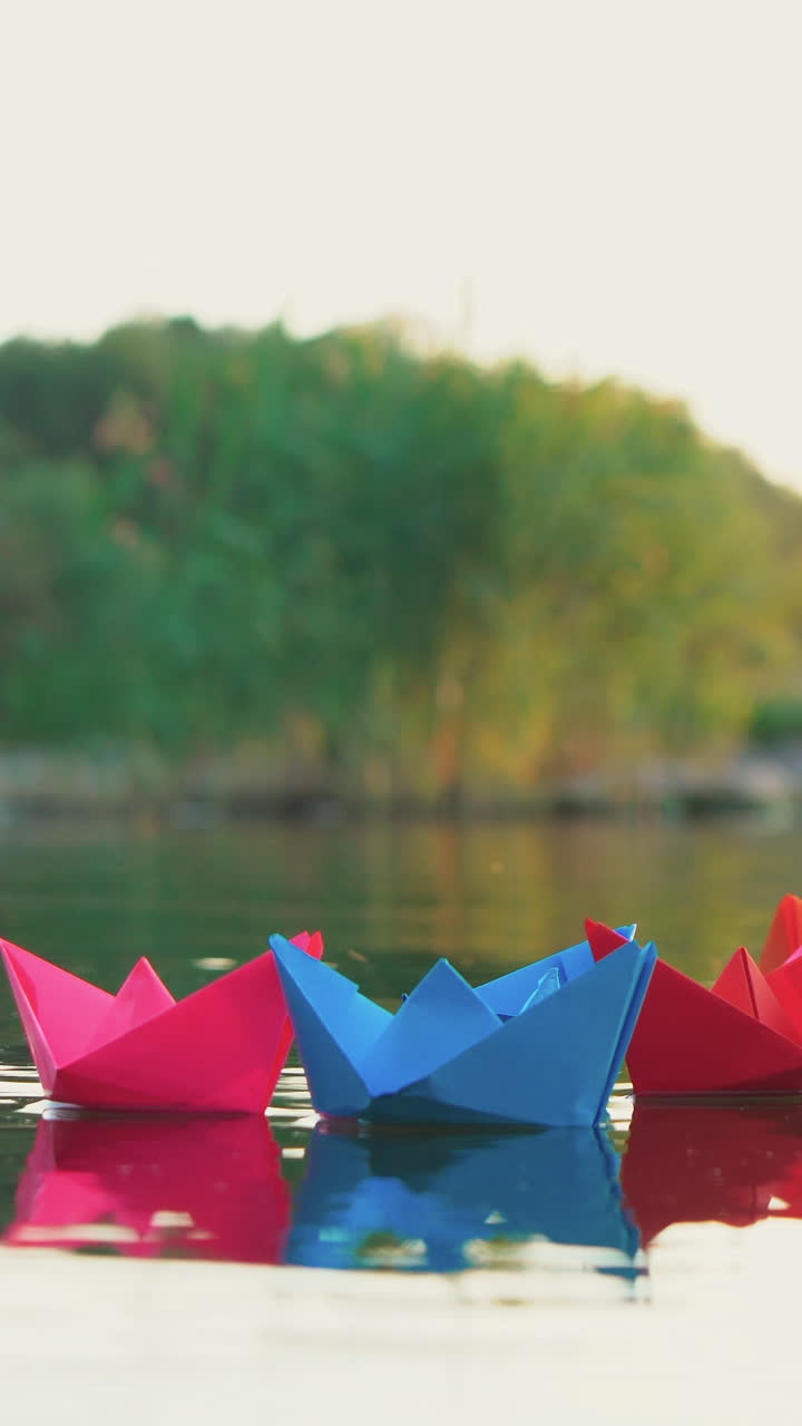 several multi-colored origami in the shape of ships are floating on the water in the lake in the evening on the background of the summer landscape. Vertical video